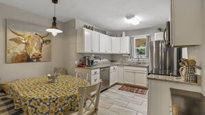 Cucina compatta con mobili bianchi, frigorifero in acciaio inossidabile e zona pranzo con tovaglia fantasia.