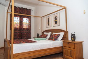 Un lit à baldaquin avec un cadre en bois, une serviette verte et des coussins à motifs. Deux cadres photo sont accrochés au mur, et une table de chevet est surmontée d’une lanterne.