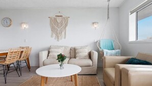 A living room with a white sofa, a beige sofa, a round coffee table, wicker chairs, a macrame wall hanging, and a hanging chair. - Maroochy Sands (Sunshine Coast)