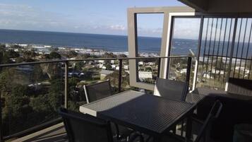 Un balcón con mesa y sillas, con vista al mar y casas a lo largo de la costa.