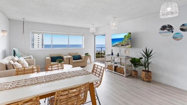 A spacious living area with a large window overlooking the sea, a dining table with wicker chairs, and a cozy seating area with cushions. - Maroochy Sands (Sunshine Coast)
