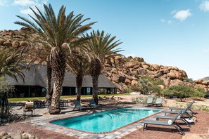A pool area with lounge chairs, palm trees, and a building with a thatched roof. - Ai Aiba The Rockpainting Lodge (Omaruru)