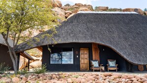 A thatched-roof building with a wooden door and windows, surrounded by rocks and a tree. - Ai Aiba The Rockpainting Lodge (Omaruru)