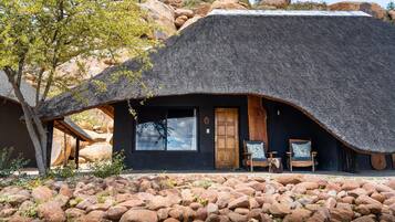 A thatched-roof building with a wooden door and windows, surrounded by rocks and a tree.