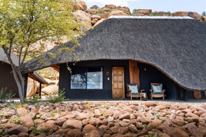 A thatched-roof building with a wooden door and windows, surrounded by rocks and a tree.