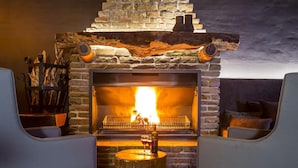 A cozy fireplace with a stone surround, a wooden log shelf, and two chairs facing the fire. - Ai Aiba The Rockpainting Lodge (Omaruru)