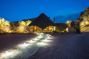 A well-lit pathway leading to a building with a thatched roof, surrounded by landscaped gardens and a rocky hillside. - Ai Aiba The Rockpainting Lodge (Omaruru)