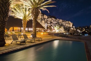 A nighttime pool area with lounge chairs, a fire pit, and a building with a rock facade. - Ai Aiba The Rockpainting Lodge (Omaruru)