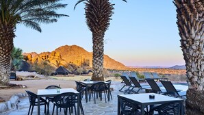 An outdoor dining area with stone flooring and black chairs, overlooking a desert landscape with mountains and a pool. - Ai Aiba The Rockpainting Lodge (Omaruru)