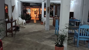 Um espaço interno amplo com piso de concreto, paredes brancas e teto de madeira. Há diversas plantas em vasos, cadeiras de plástico branco e uma cadeira azul. Uma porta dá acesso a outro cômodo com uma mesa e cadeiras.