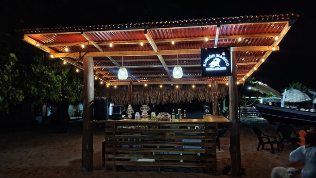 Um bar à beira-mar com estrutura de madeira, uma tela de TV e diversos copos e garrafas no balcão.