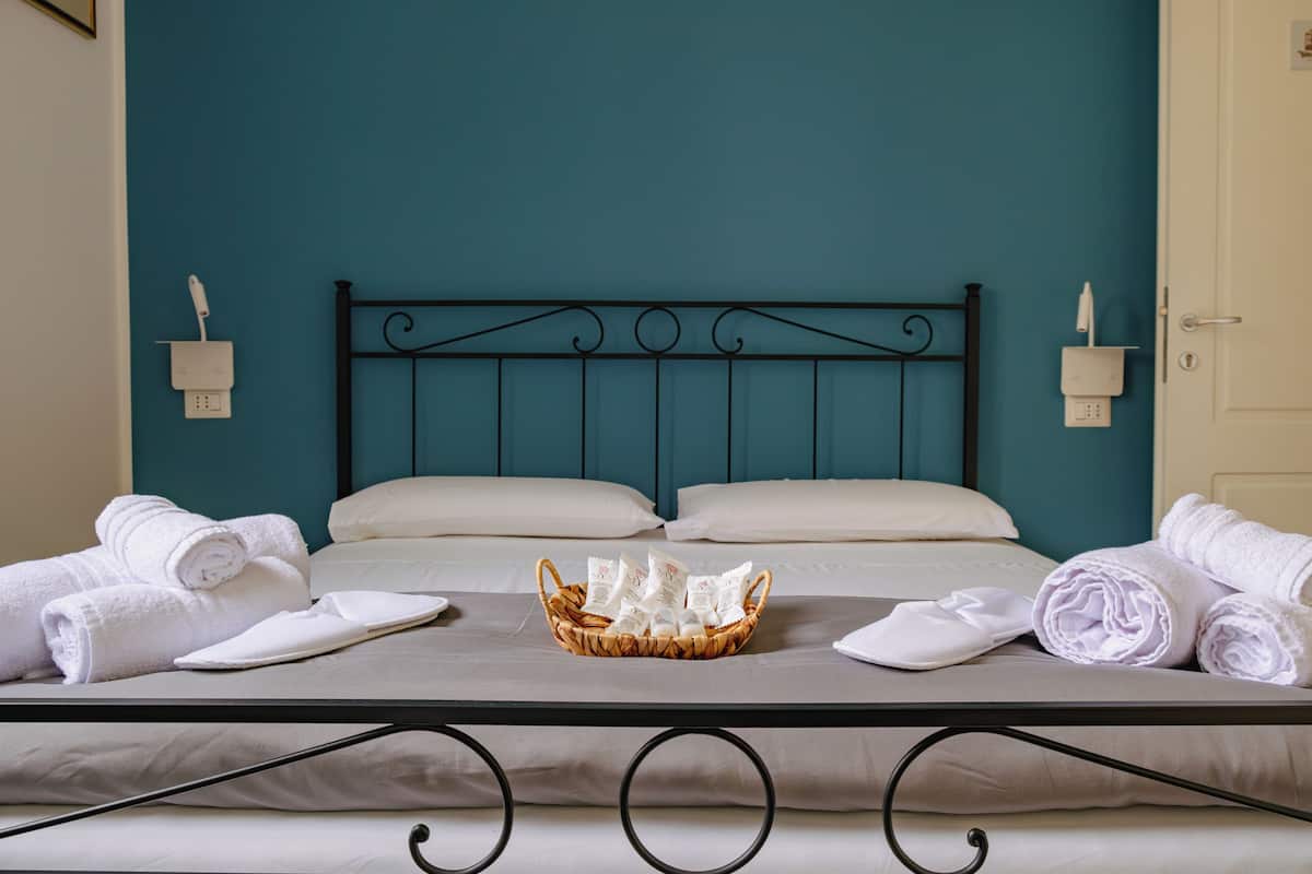 Un lit bien fait, avec du linge de lit blanc et un panier contenant des articles de toilette, sur un lit à structure métallique dans une pièce aux murs de couleur turquoise.