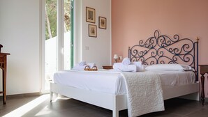 A neatly made bed with white linens and a decorative headboard, a bedside table with a lamp, and a window with natural light. - Villa Rubino Guest house Mondello (Palermo)