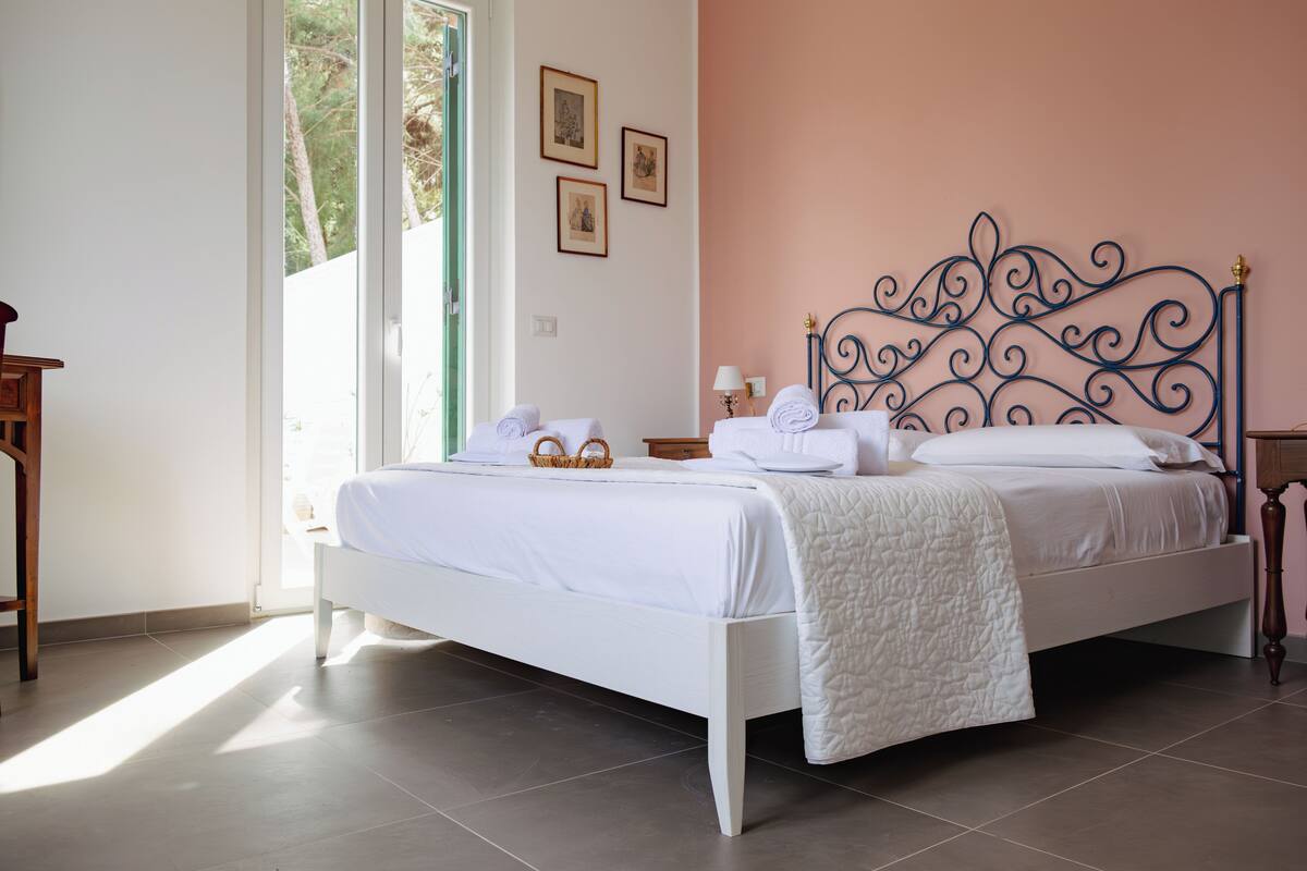 Un lit bien fait, avec du linge de lit blanc et une tête de lit décorative, une table de chevet avec une lampe et une fenêtre laissant entrer la lumière naturelle.