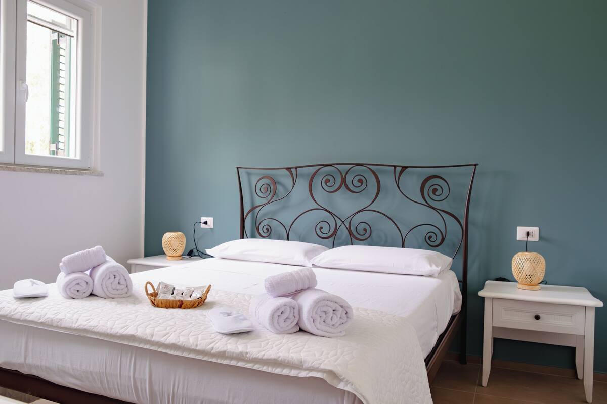 Un lit bien fait, avec des serviettes pliées et un panier contenant des articles de toilette, une lampe de chevet et une fenêtre avec des stores.