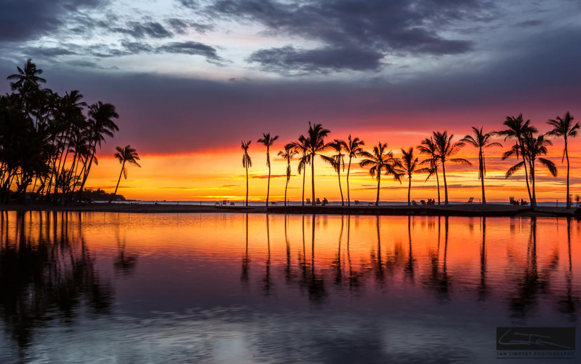 Een zonsondergang boven een rustig strand, met palmbomen die in het water weerspiegeld worden.