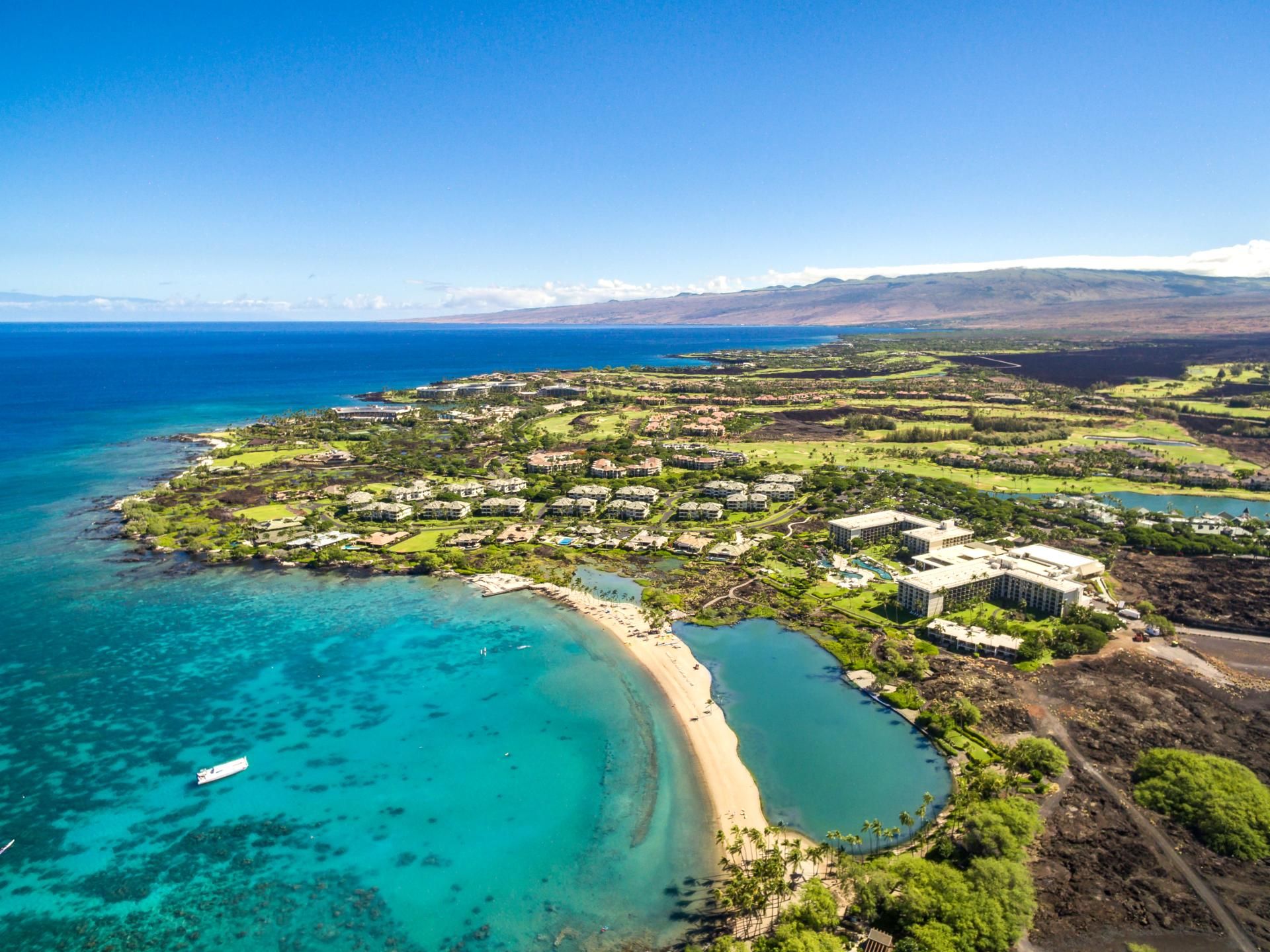 An aerial view of a coastal resort with a beach, clear turquoise waters, and a mix of residential and commercial buildings.