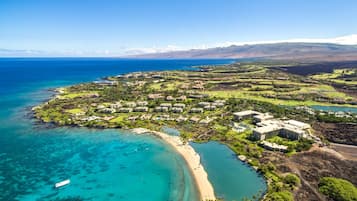 An aerial view of a coastal resort with a beach, clear turquoise waters, and a mix of residential and commercial buildings.