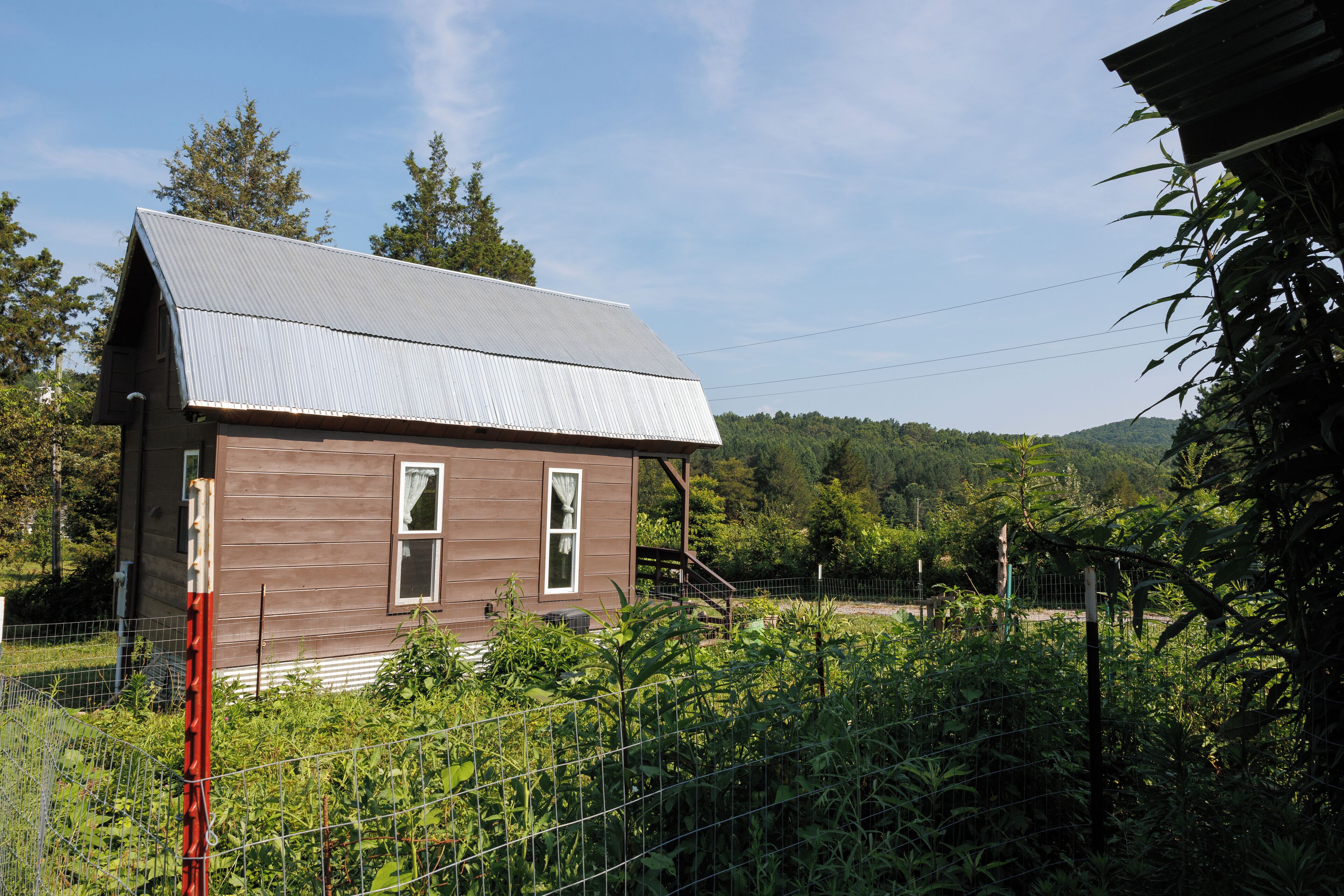 A small, single-story house with a sloped metal roof, surrounded by dense vegetation and a wire fence.