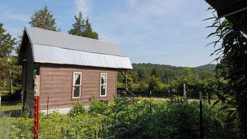 A small, single-story house with a sloped metal roof, surrounded by dense vegetation and a wire fence.
