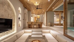 A modern living room with a large TV, comfortable seating, a dining area, and a view of a bedroom and pool.