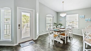 A dining area with a table set for four, white chairs, a chandelier, and a view of the outdoors through large windows.