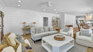 Un salón moderno con un sofá blanco, un sillón blanco y una mesita de centro. Se aprecia una estantería y un comedor al fondo.