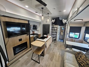 A compact RV kitchen with a stove, microwave, and a dining area with a bench. - Spacious 2025 Destination RV Sleeps 9 (Lorena)