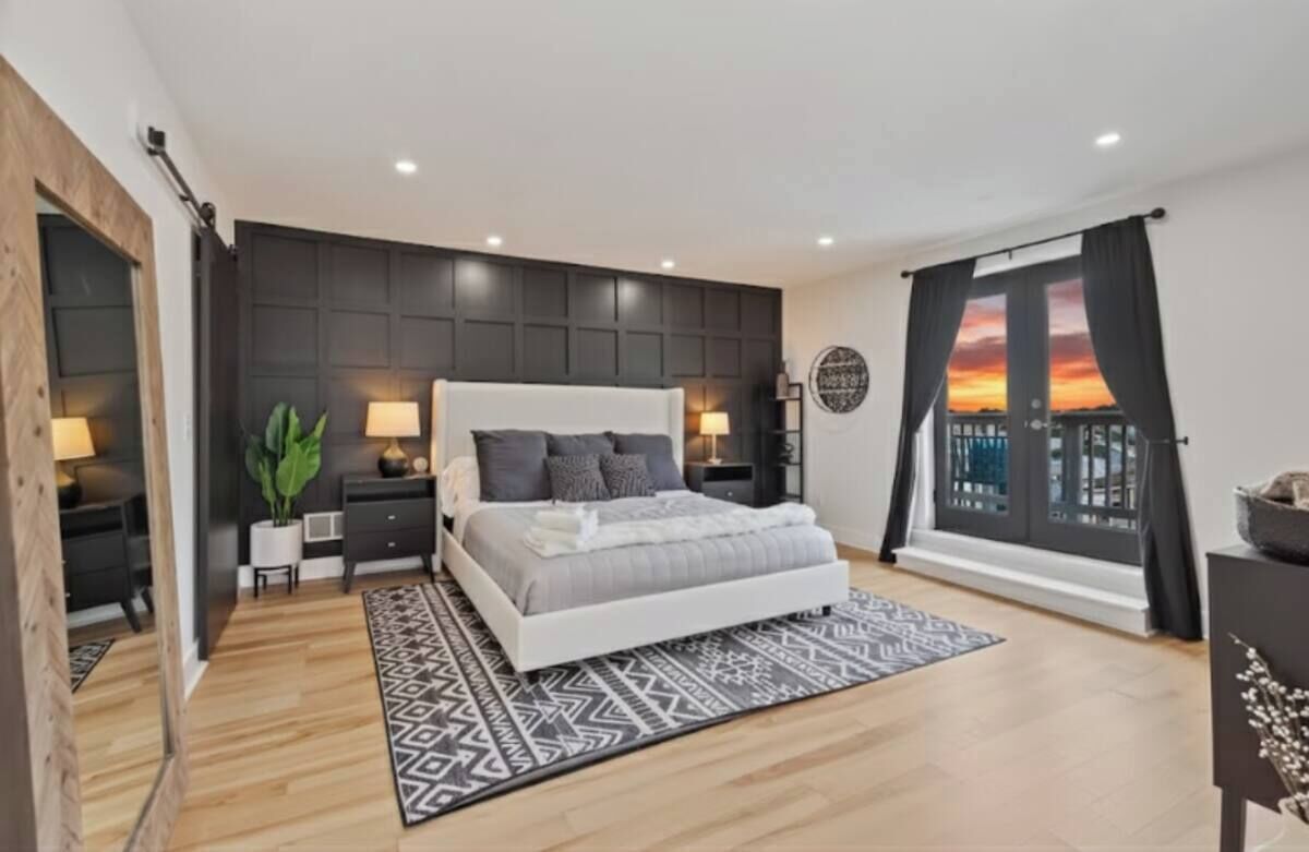 Een moderne slaapkamer met een groot bed, nachtkastjes, een plant in pot en uitzicht op een ondergaande zon door een raam.