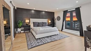 A modern bedroom with a large bed, bedside tables, a nightstand, a potted plant, and a view of a sunset through a window. - 3BR Stylish Retreat, Minutes from Downtown Pittsburgh, Free 3-Car Parking (Pittsburgh)