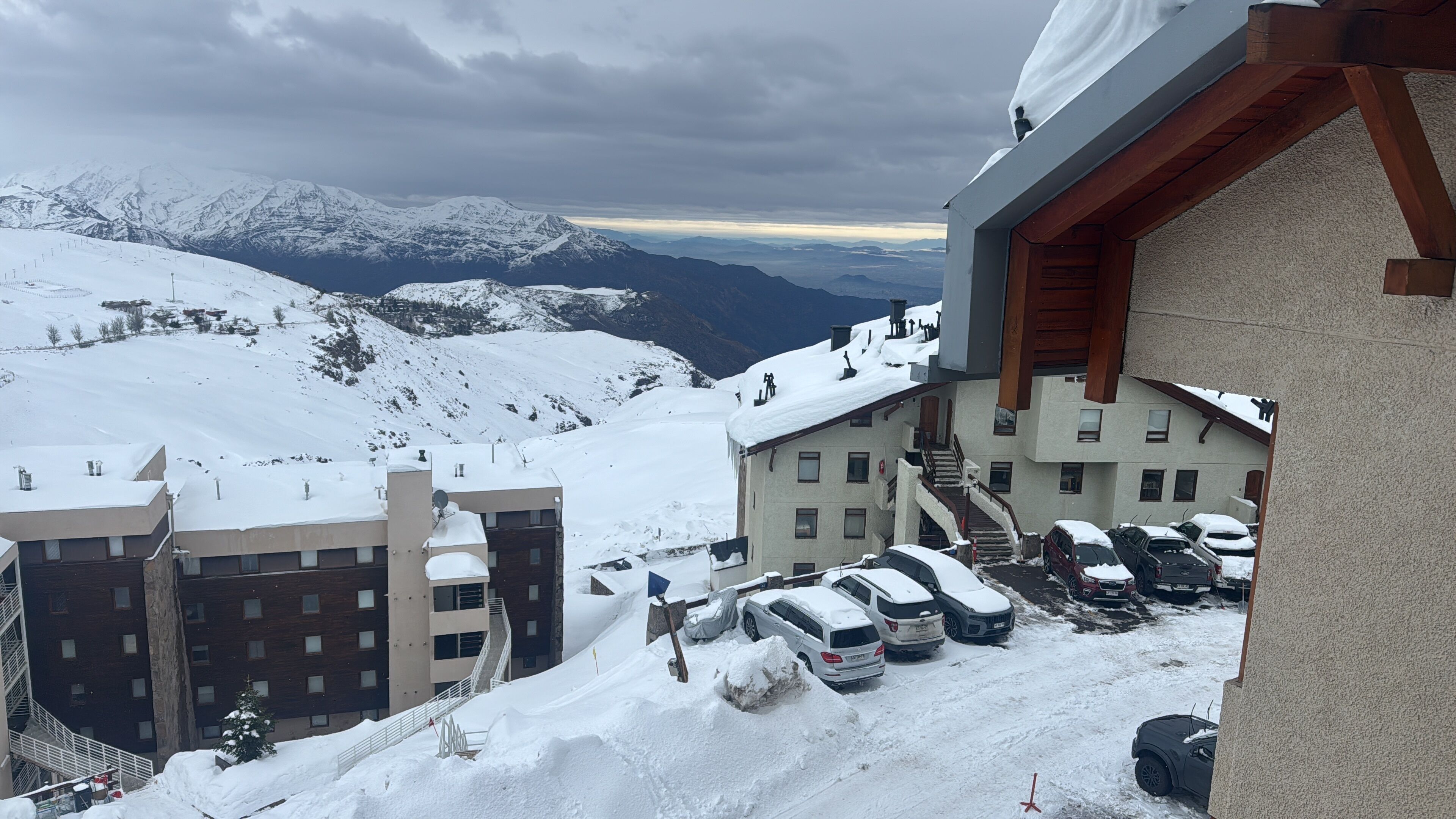Un complejo turístico en la montaña cubierto de nieve, con varios edificios, un estacionamiento y vistas a montañas distantes.
