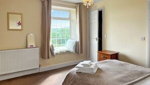 Quarto com cama grande, janela com cortinas, radiador, mesa de cabeceira de madeira e porta.