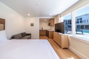 A modern hotel room with a large bed, a sofa, a TV, a kitchenette, and a view of a pool. - Ocean's Edge Luxury Rentals (Ocean City)