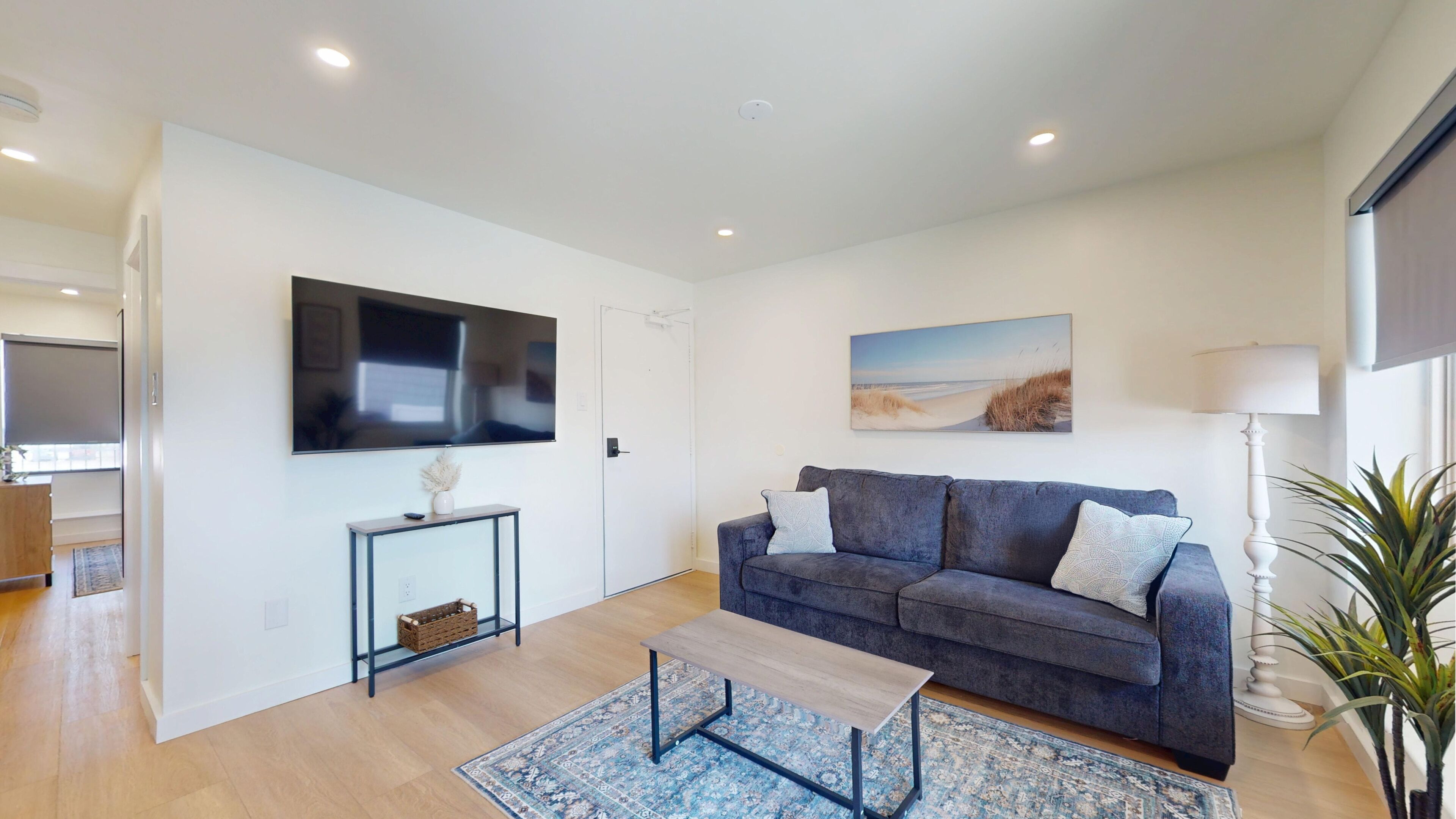 Una sala de estar con un televisor de pantalla plana, un sofá gris, una mesa de centro, un tapete estampado y un cuadro de una playa.