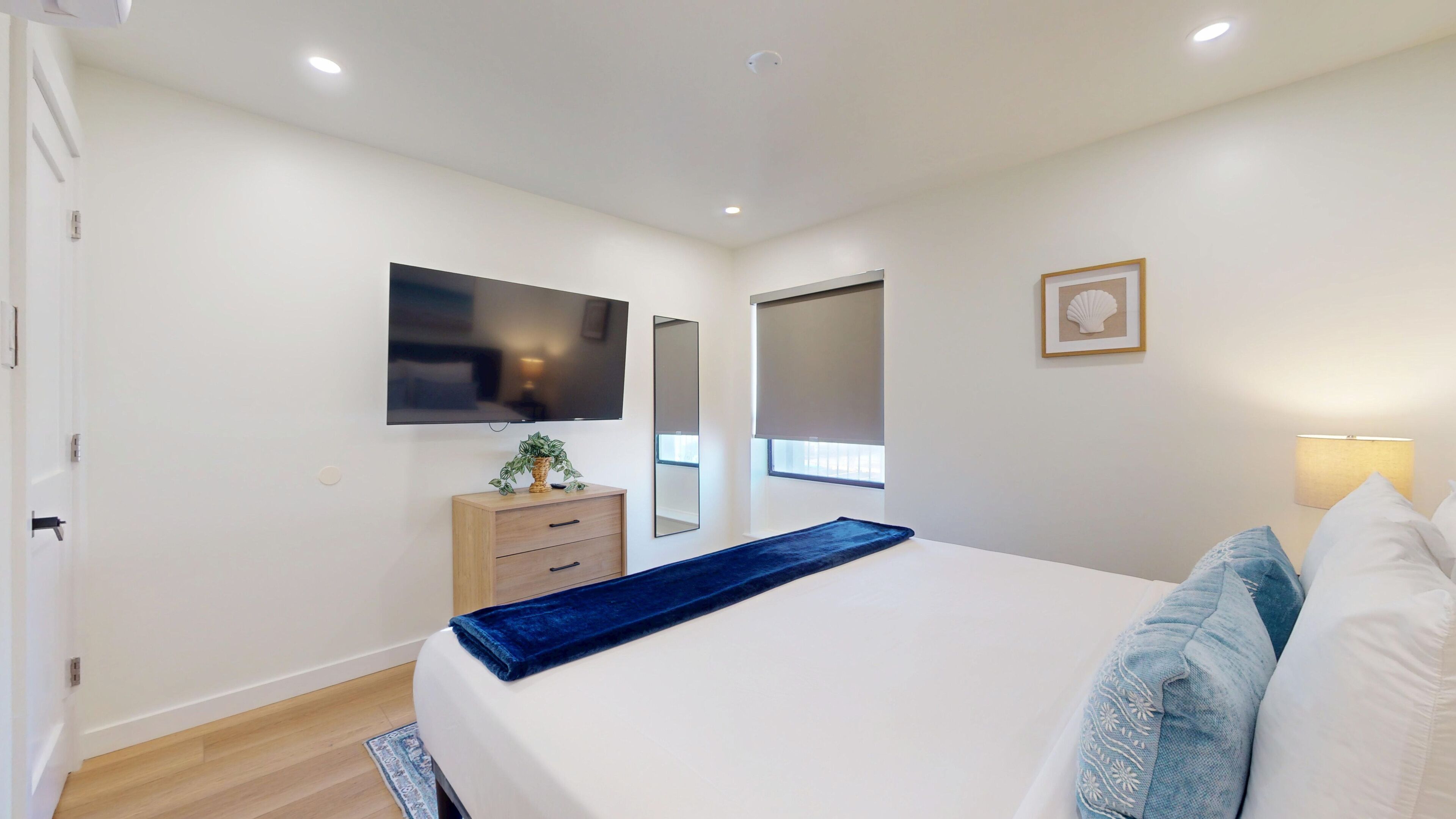 A bedroom with a bed, a TV, a dresser, a mirror, a window with a blind, and a wall-mounted lamp.