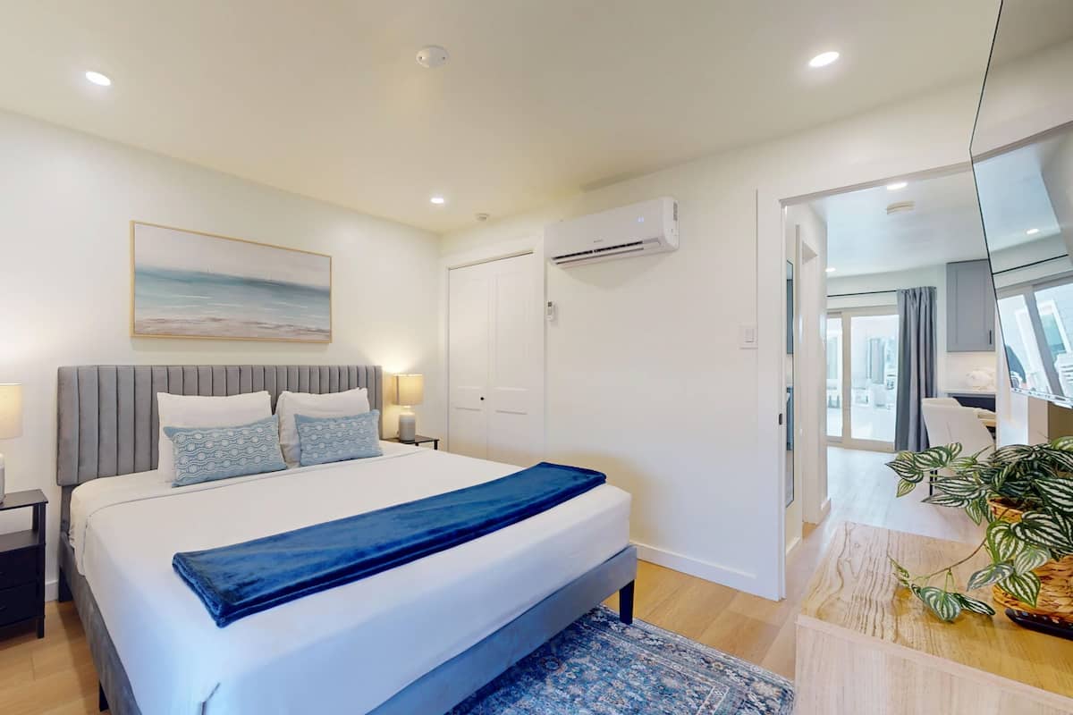 A bedroom with a bed, bedside tables, a nightstand, a painting, an air conditioning unit, and a view to the outside.