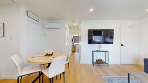 A modern living room with a round dining table, white chairs, a flat-screen TV mounted on the wall, and a small console table with a vase and remote control.