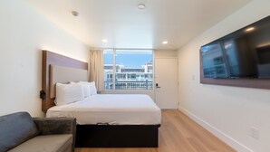 Une chambre d’hôtel moderne équipée d’un lit, d’un canapé, d’une télévision et d’une grande fenêtre donnant sur des immeubles.