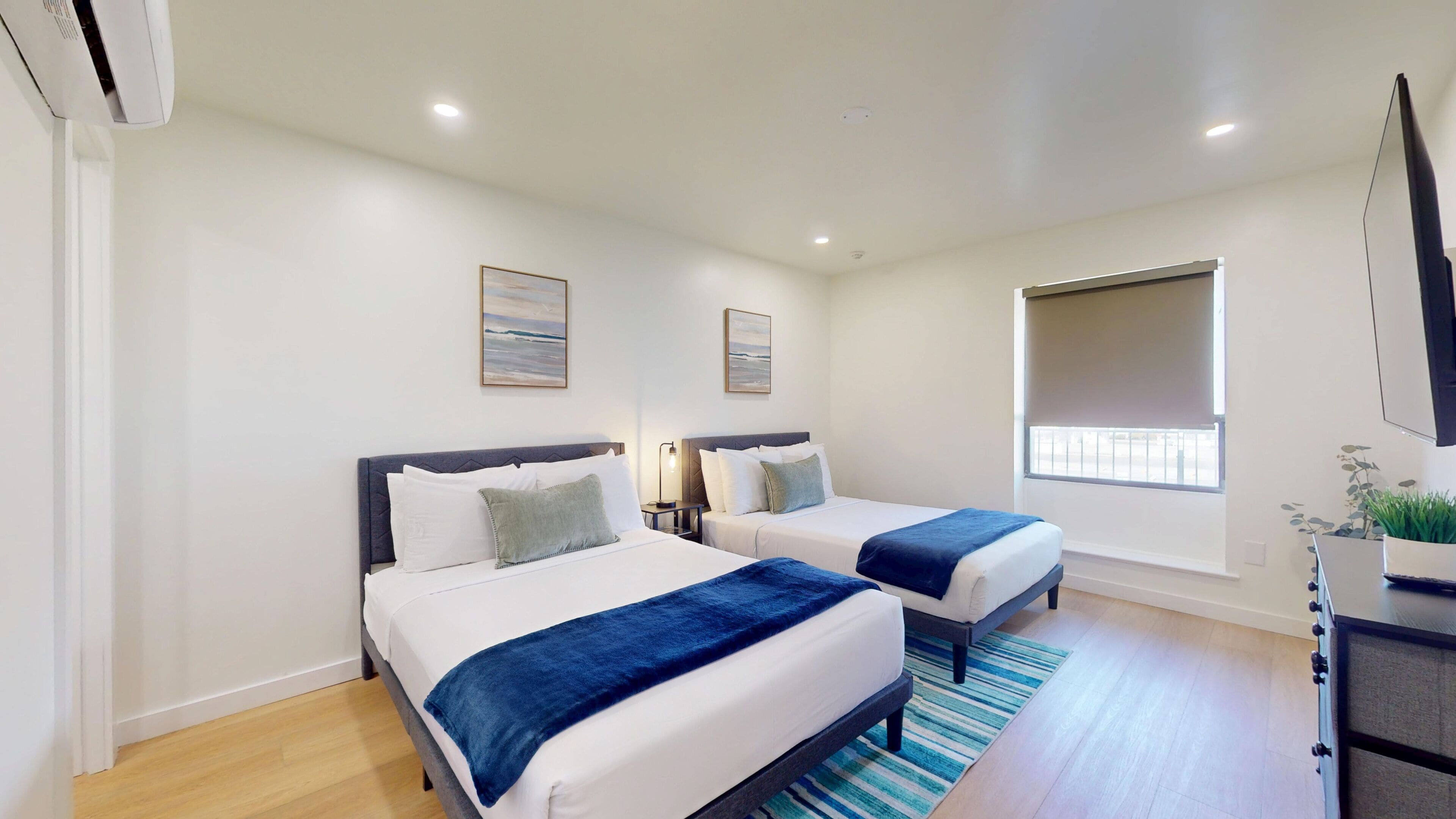 A hotel room with two beds, a window, a flat-screen TV, and a ceiling-mounted air conditioner.