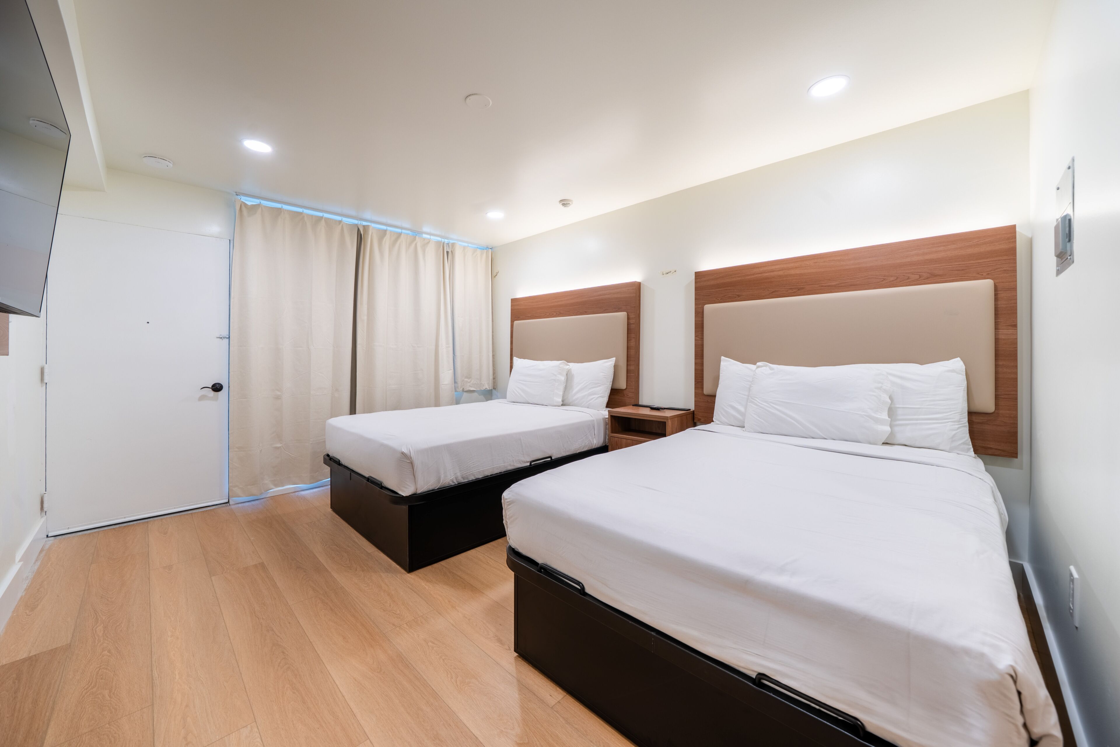 Quarto de hotel com duas camas, cabeceira de madeira, porta branca e cortina em tom claro.