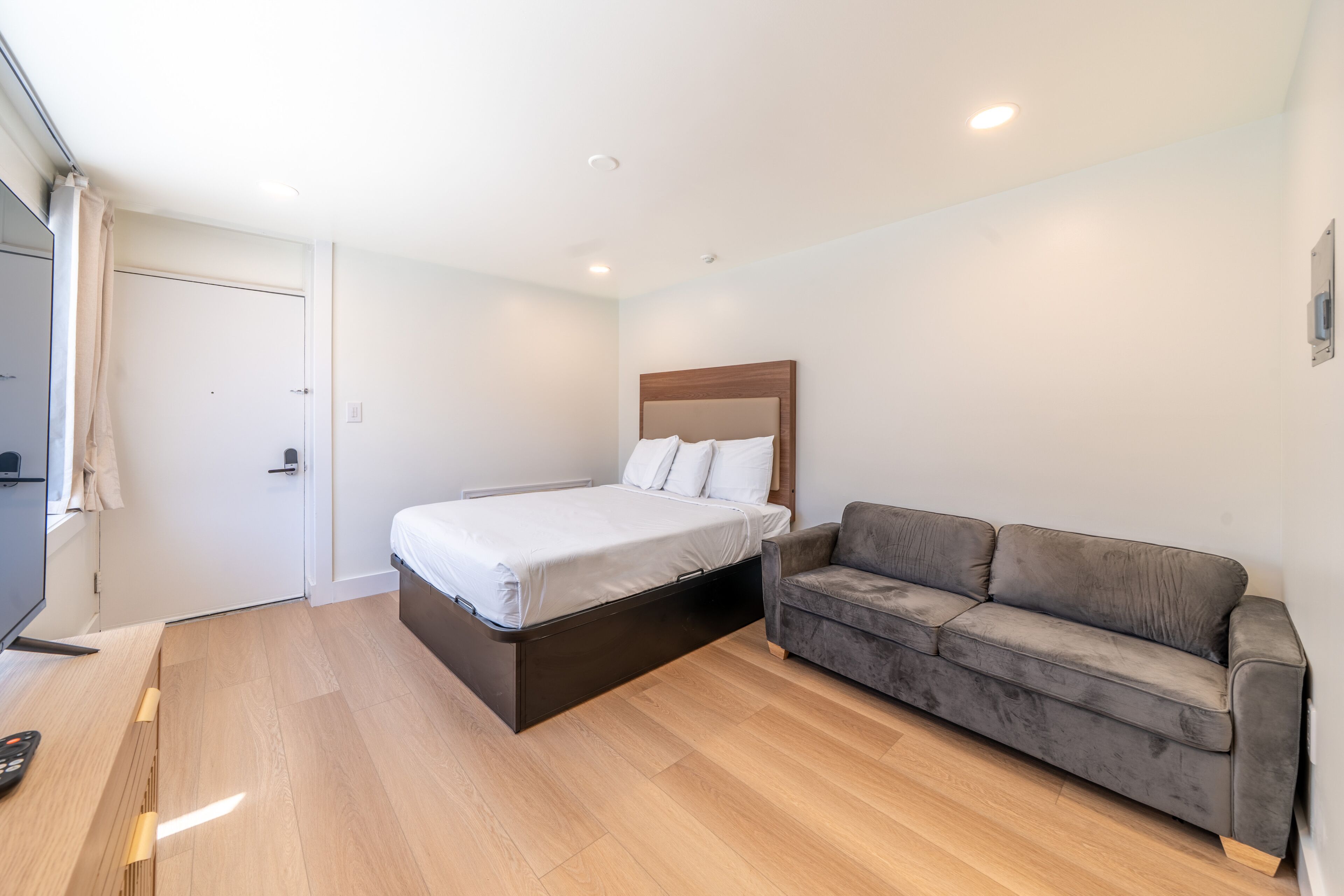 A bedroom with a bed, a gray sofa, and a wooden floor.