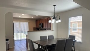Un comedor con una mesa rectangular y seis sillas, una cocina con armarios de madera y un ventanal con persianas.