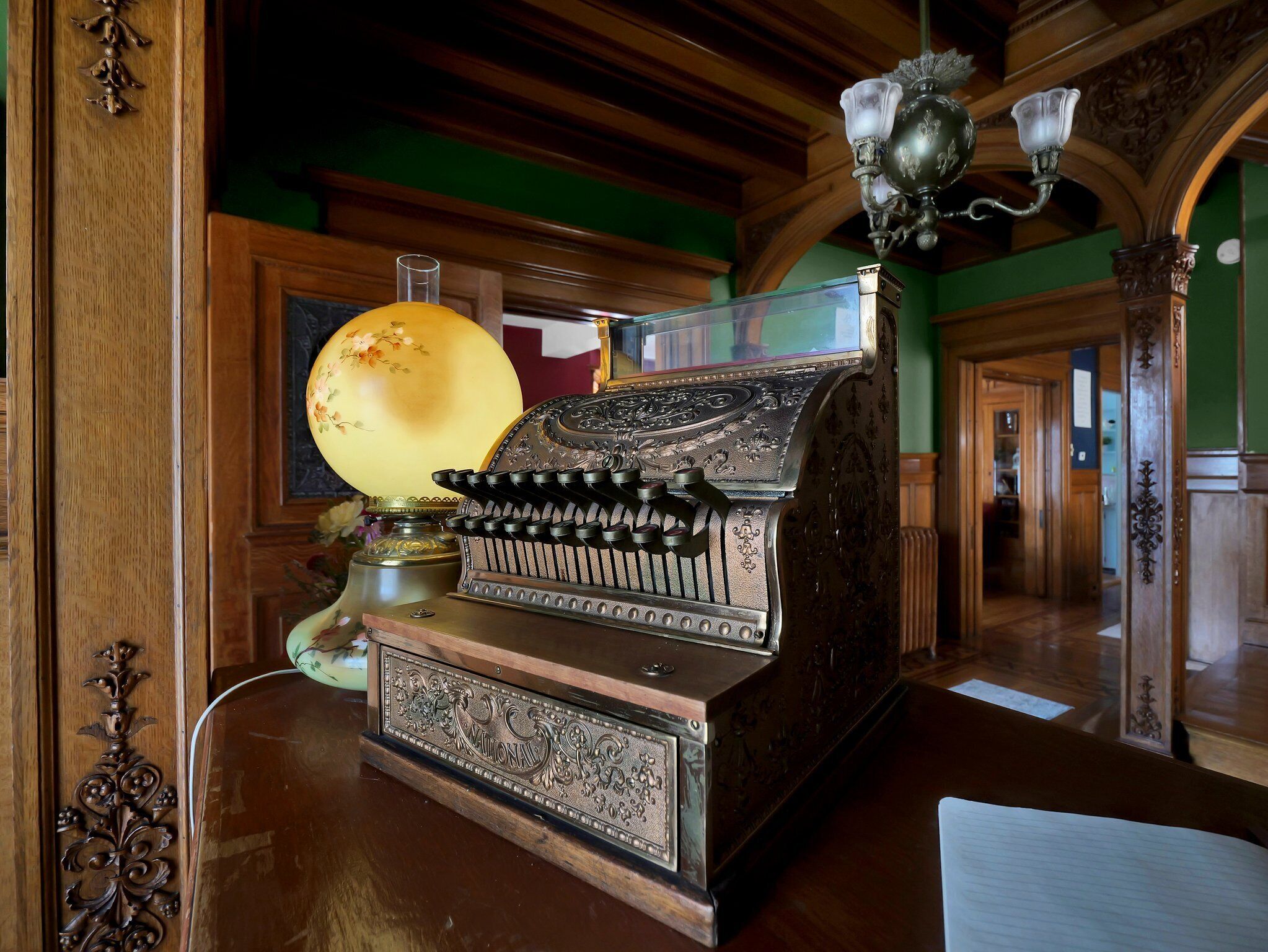 A vintage typewriter with ornate designs, a lamp with floral patterns, and a chandelier in a room with wooden paneling and green walls.