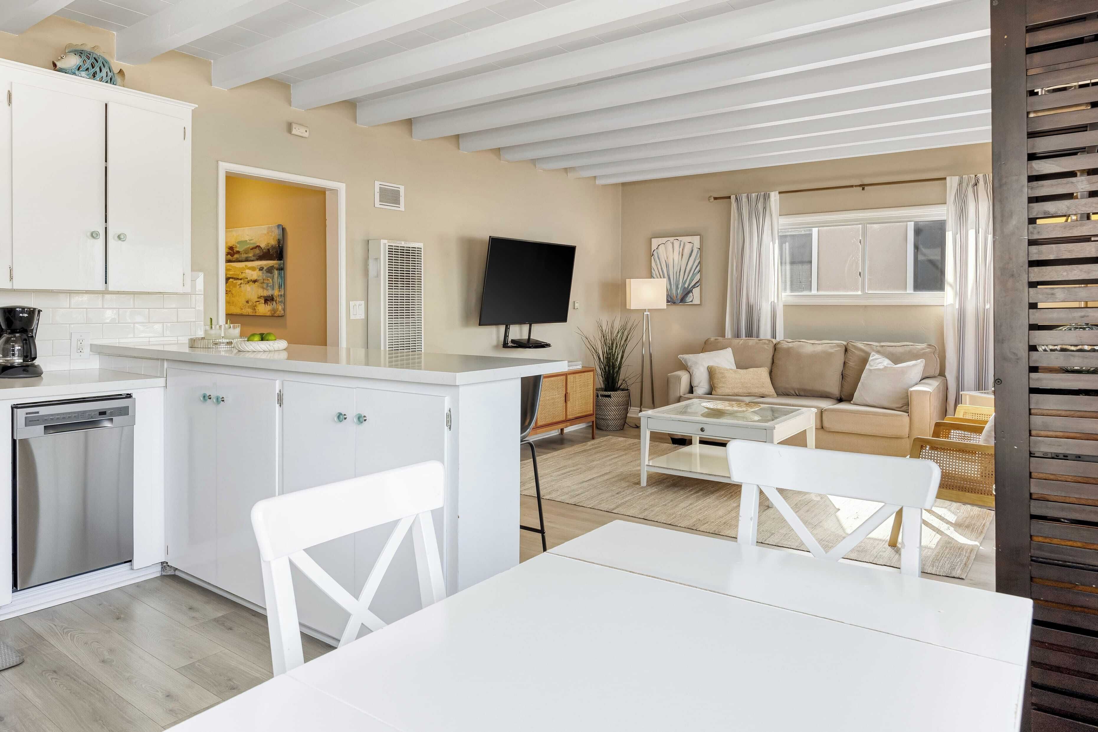 A modern kitchen with white cabinets, a dishwasher, and a dining area with a white table and chairs. There is a sofa and a coffee table in the living area.