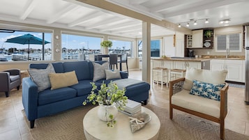 Un salón espacioso con un sofá azul, una silla de madera y una mesa de centro con adornos. Un ventanal grande ofrece vistas a un puerto deportivo.