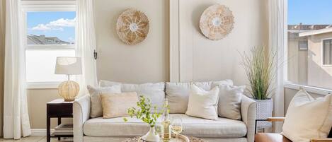 A living room with a beige sofa, a round coffee table with decorative items, a side table with a lamp, and two wall-mounted decorative pieces.