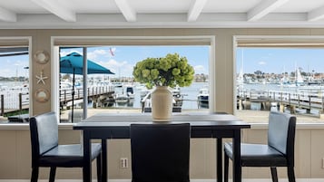 Un comedor con una mesa y sillas, un florero con flores y vistas a un puerto deportivo con barcos y un puente.