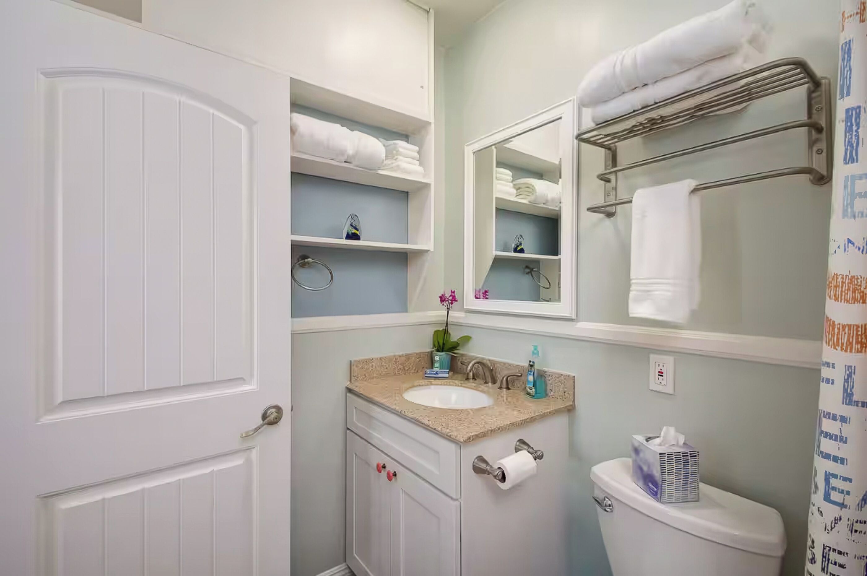 A bathroom with a white door, a sink with a countertop, a mirror above the sink, a towel rack, and a toilet.