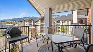 A balcony with a grill, chairs, and a table, overlooking a residential area with mountains in the background.