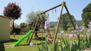 A wooden swing set, a green slide, and purple flowers in a backyard.
